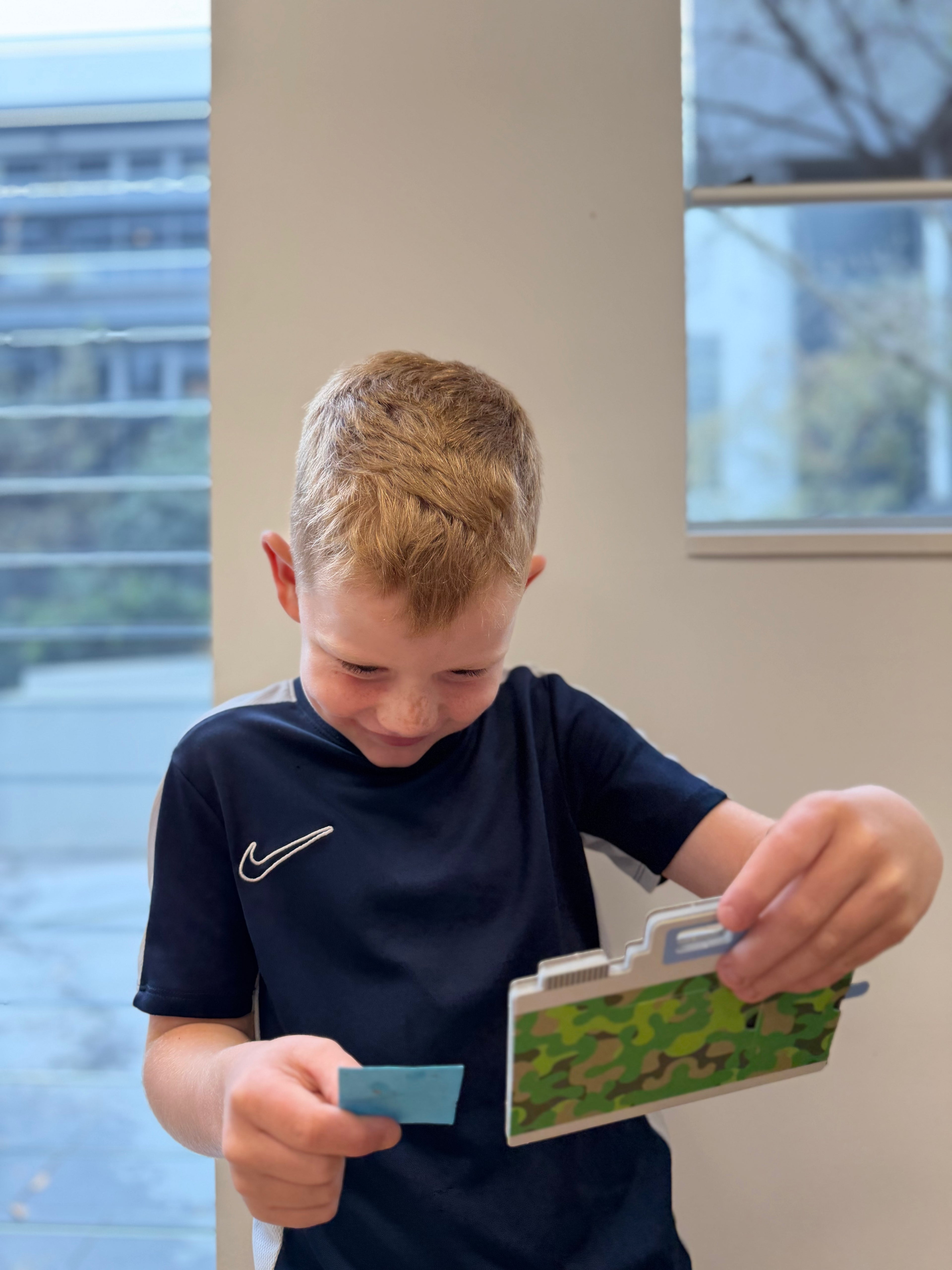 A young boy with blond hair wearing a navy blue t-shirt, looking down at a small green camouflage camera he is holding while holding cyanotype paper in his other hand, in an indoor setting.