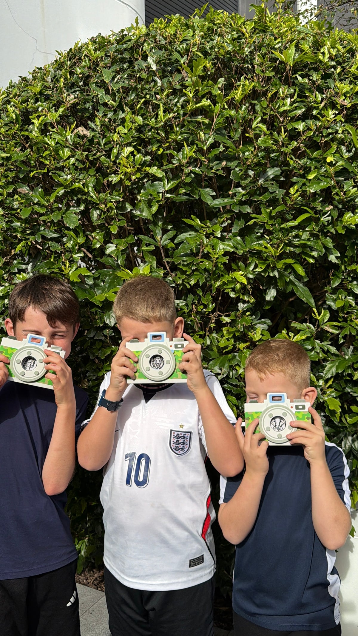 Three boys standing in front of a green hedge, each holding a green and white toy camera in front of their face.
